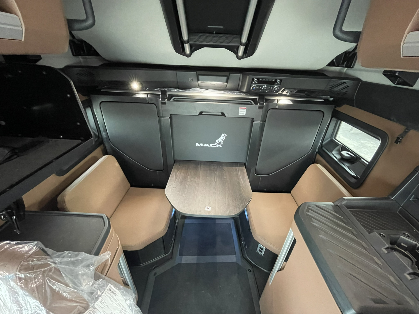 Interior of a new Mack PR64T76H sleeper cab showing tan seats, table, and control panels. Text visible: 'MACK' on the rear console; no other legible labels.