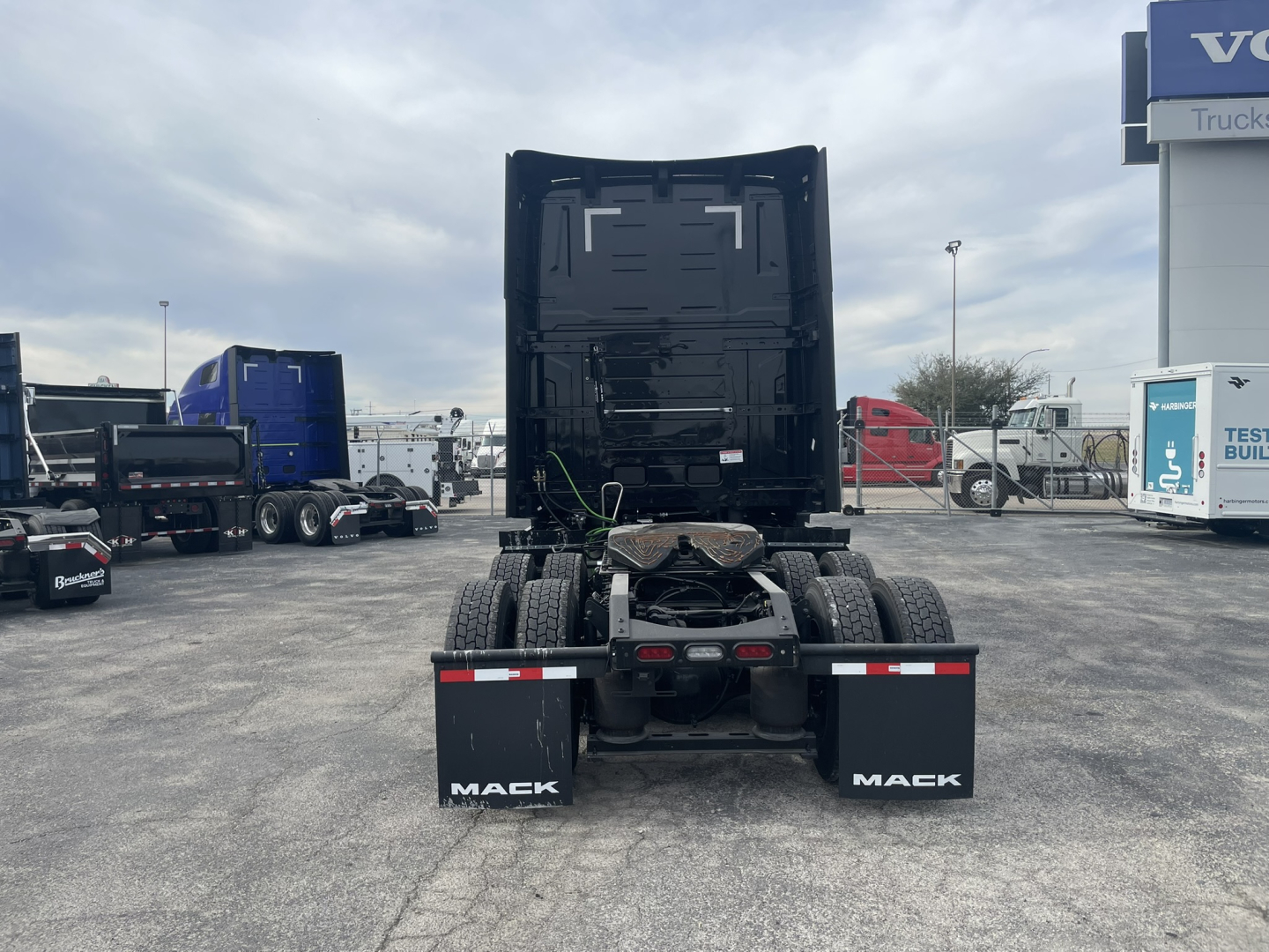 Rear view of a black Mack PR64T76H semi-truck tractor with fifth wheel and dual rear axles, mud flaps labeled 'MAEK' (MACK misspelling) visible; surrounding trucks in a dealership lot.