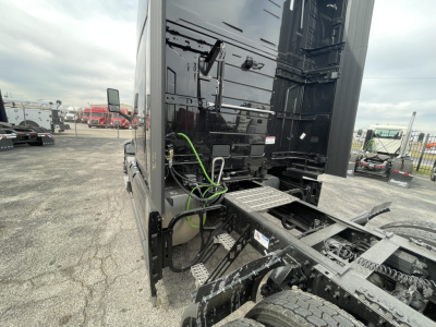 Truck cab and chassis area of a 2026 Mack PR64T76H with visible rear engine and side steps, black paint, green air hoses, and metal frame components.