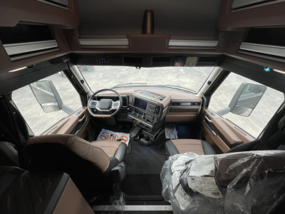 Truck cab interior with steering wheel, dashboard, and seats visible; plastic-wrapped seat on passenger side; overhead storage; center console with controls.