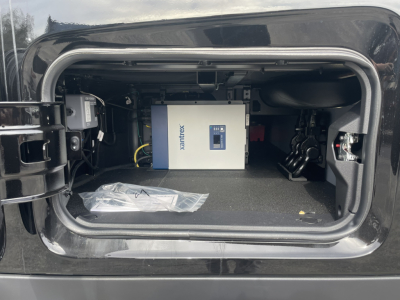 Truck storage compartment with an Enovix/Box-like electrical unit and cables inside, visible label 'Maximex' or similar on the blue/white device; compact space with carpeting and a bag of parts.