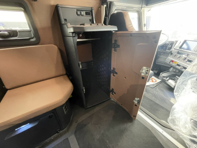 Truck cab interior with open cabinet storage in sleeper area; tan upholstery; driver seat and dashboard visible; open vertical cabinet with metal hinges.