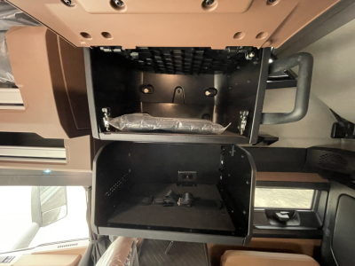 Cab interior of a Mack PR64T76H showing an open overhead storage compartment and lower glovebox with a wrapped item inside.