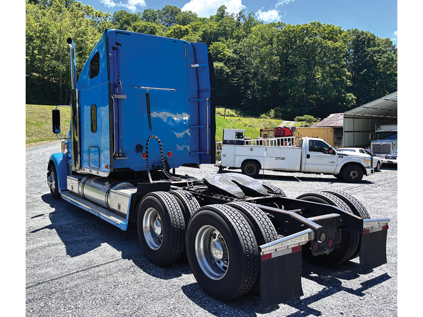 Freightliner Coronado tractor cab in blue with dual rear axles and fifth wheel, viewed from rear-left; includes fuel tanks, exhaust, and parking lot in background.
