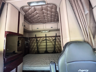 Truck sleeper cab interior showing quilted beige headliner and sleeping berth with mattress, storage nets, drawers, and beige seating.