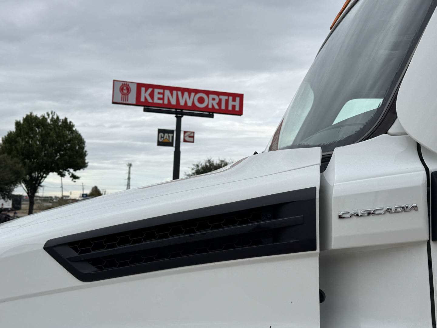 Truck cab of a white Freightliner Cascadia with a Kenworth sign in the background, exterior view from the passenger side window.