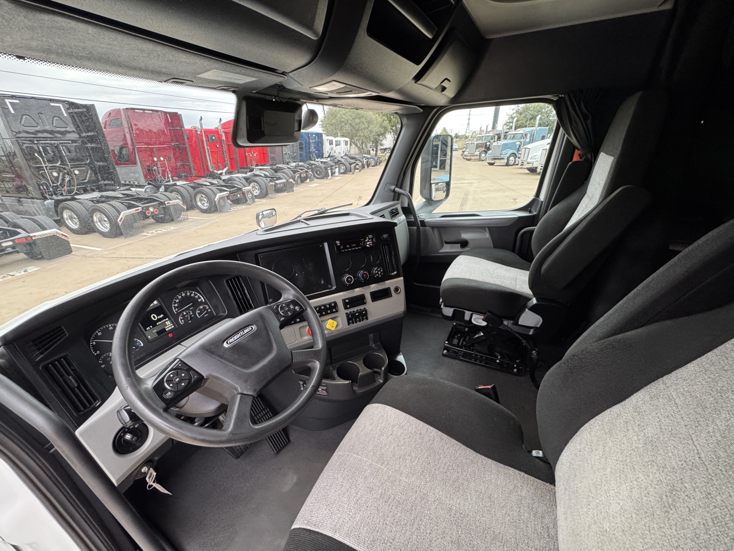 Truck cab interior of a Freightliner sleeper with steering wheel, dashboard, and driver seat visible from passenger side, components and controls in view.