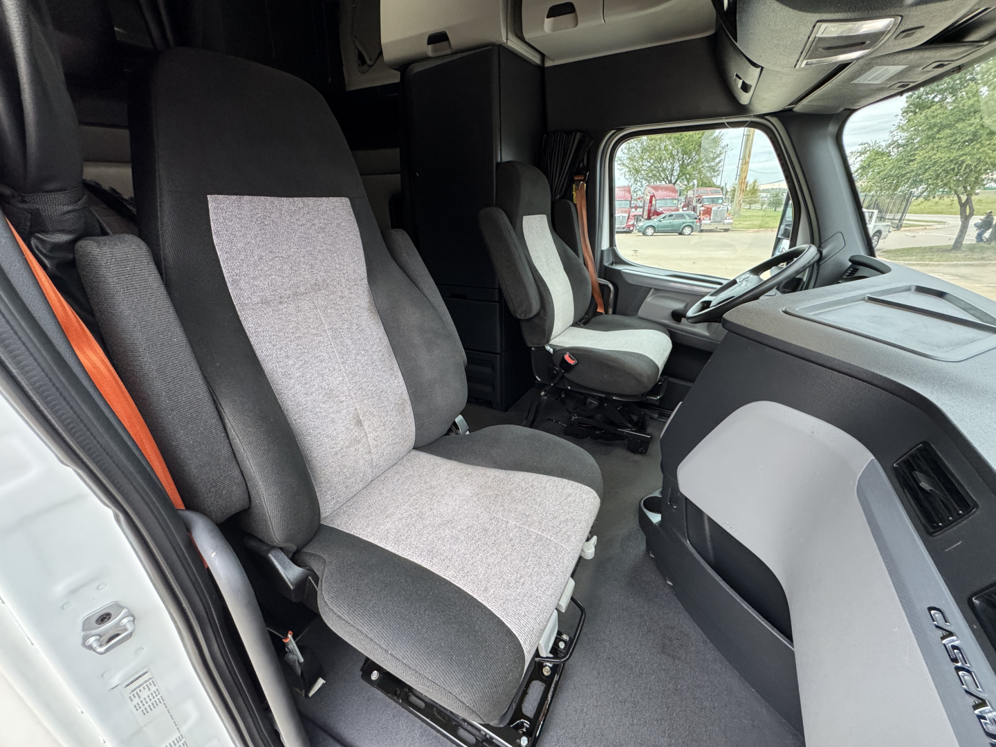 Cab interior of a Freightliner truck showing two front seats and dashboard view with steering wheel on the passenger side; fabric seats in gray tones.