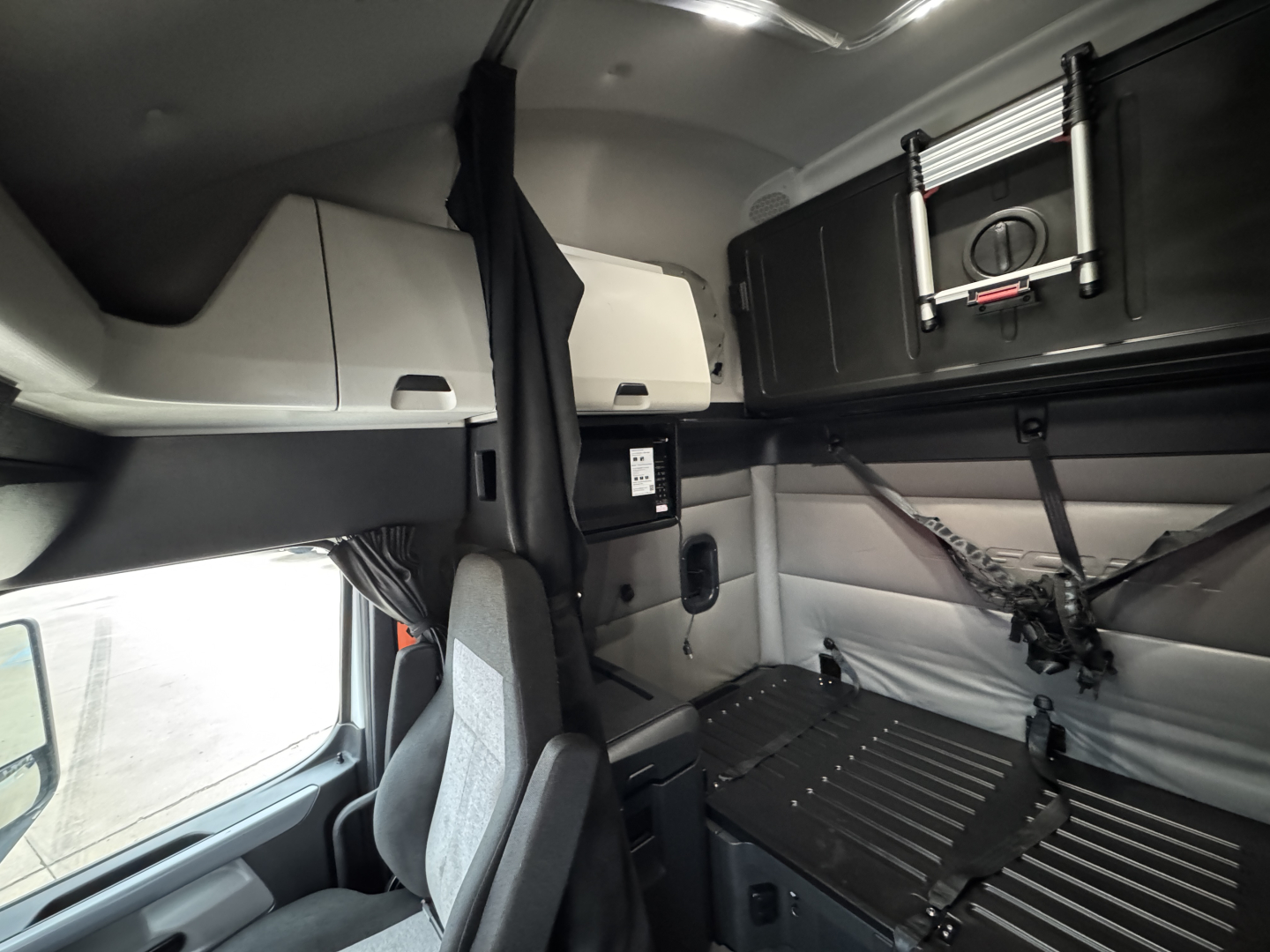 Cab interior of a Freightliner truck sleeper with storage compartments and seating area visible, including overhead cabinets and a windowed space.