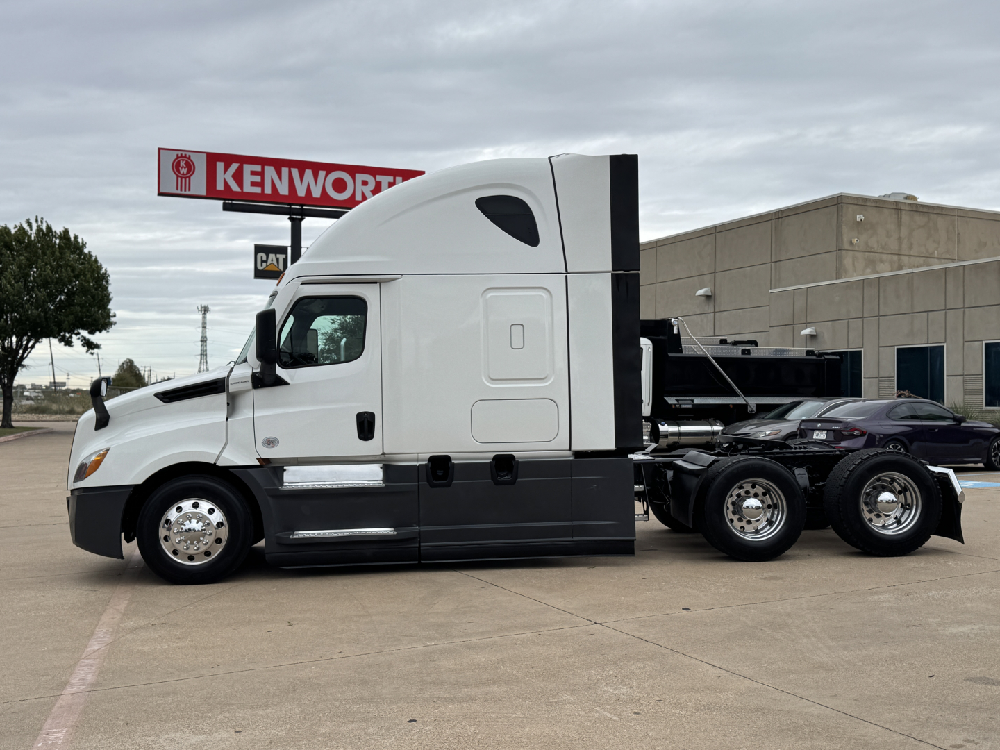 Truck side profile showing a Freightliner PT126064ST sleeper tractor chassis with dual rear axles, light gray cab, black lower body, and Kenworth sign in background.