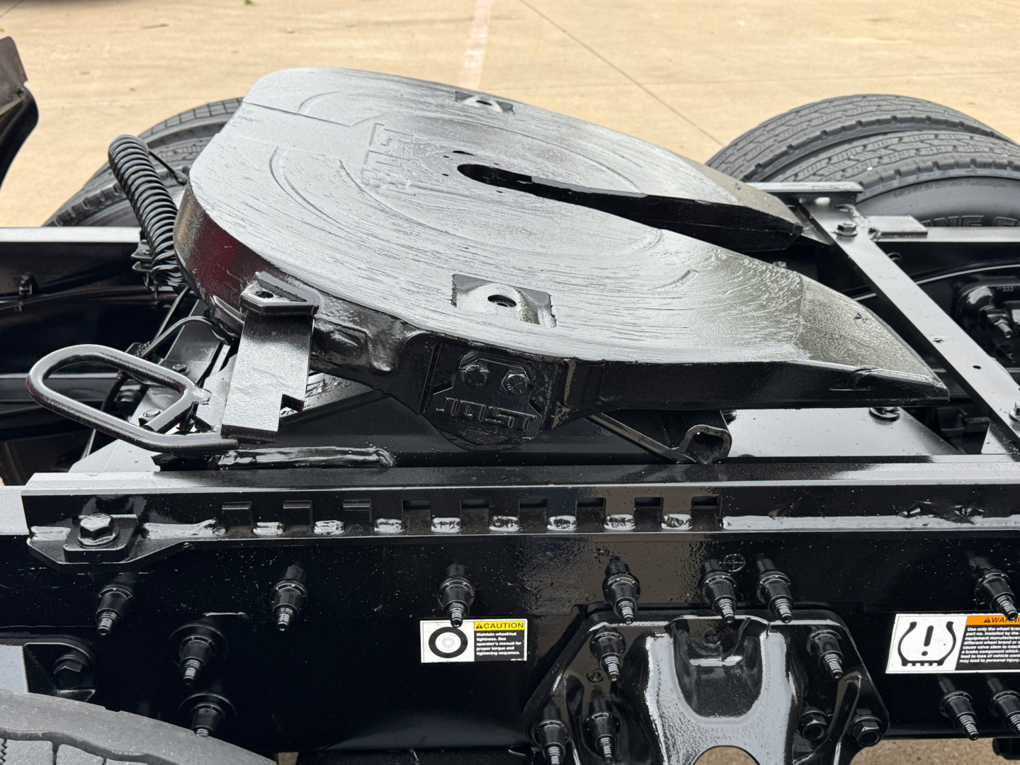 Tractor truck chassis with fifth wheel and air lines visible; circular MT-style kingpin mount on top of the frame near the cab; multiple electrical/air hoses and warning decals on the side rail.