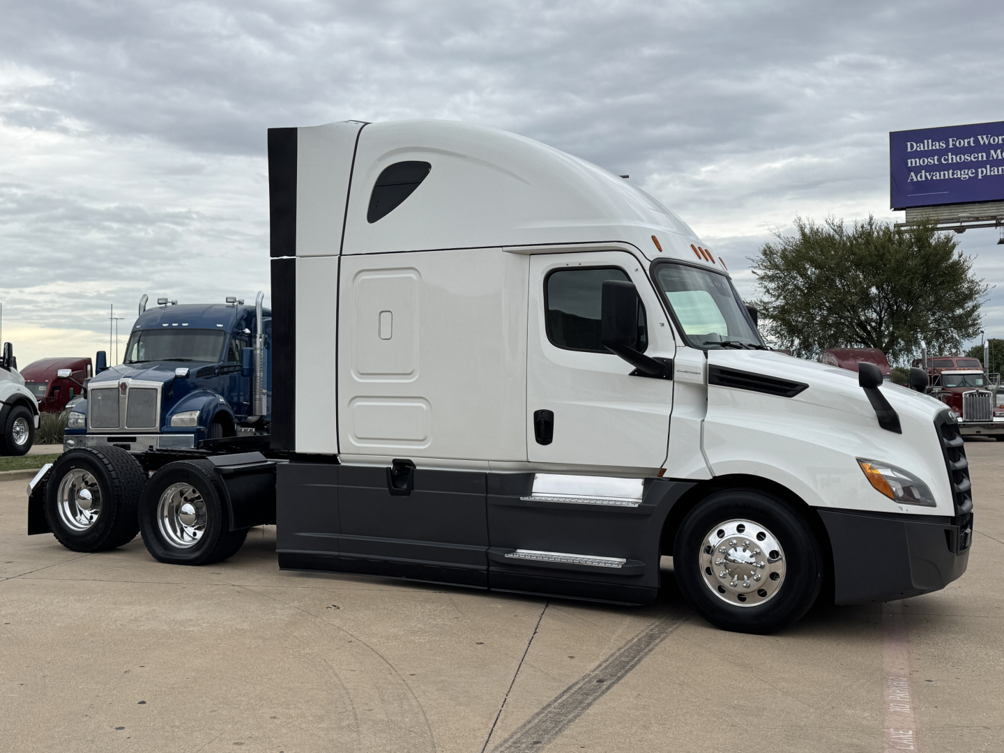 Visible side view of a white Freightliner PT126064ST heavy-duty truck, with a high sleeper cab, black lower panel, chrome wheels, and a blue truck in the background.