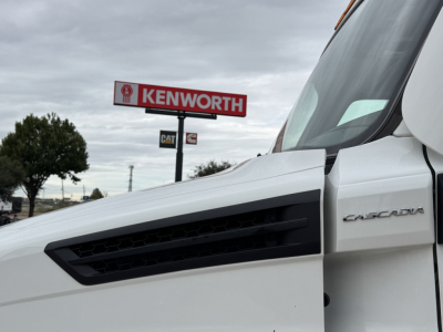 Truck cab of a white Freightliner Cascadia with a Kenworth sign in the background, exterior view from the passenger side window.