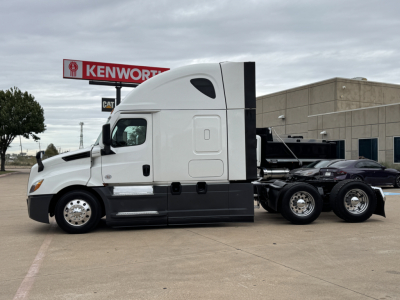 Truck side profile showing a Freightliner PT126064ST sleeper tractor chassis with dual rear axles, light gray cab, black lower body, and Kenworth sign in background.