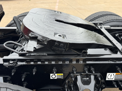 Tractor truck chassis with fifth wheel and air lines visible; circular MT-style kingpin mount on top of the frame near the cab; multiple electrical/air hoses and warning decals on the side rail.