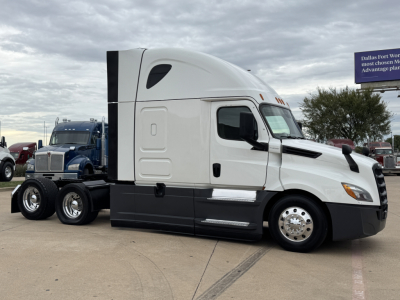 Visible side view of a white Freightliner PT126064ST heavy-duty truck, with a high sleeper cab, black lower panel, chrome wheels, and a blue truck in the background.
