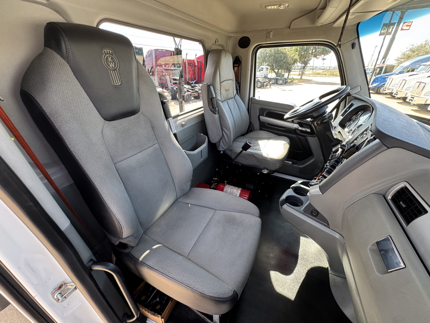 Kenworth T680 cab interior showing front seats and dashboard, light gray upholstery, steering wheel, and center console.