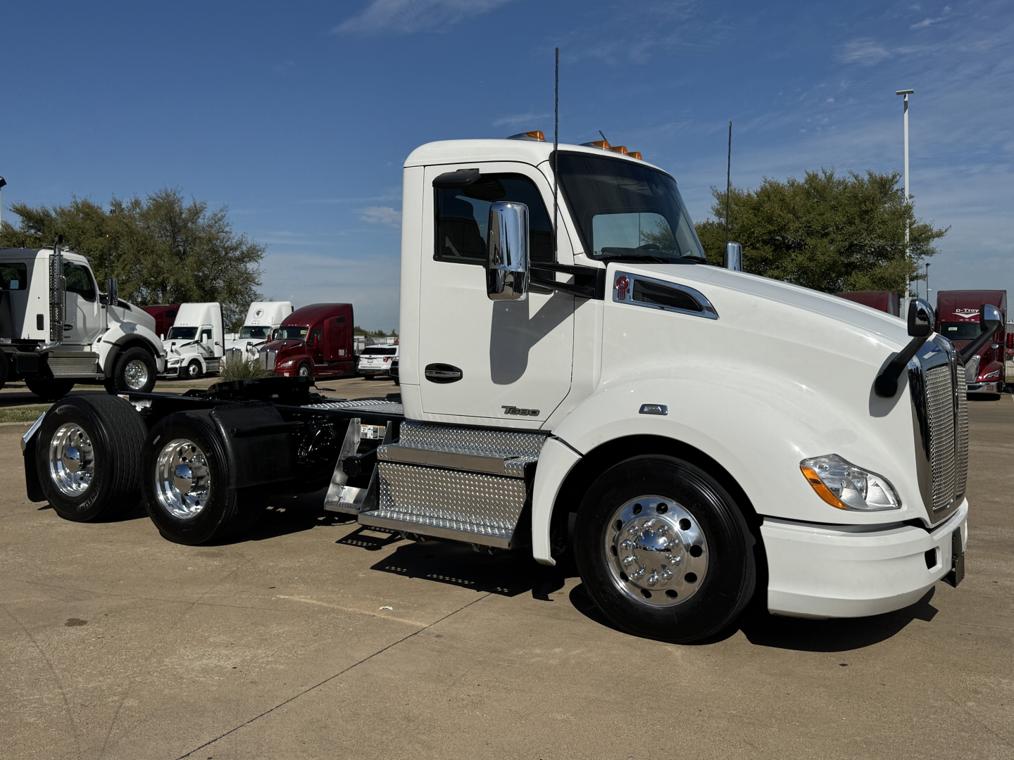 Kenworth T680 conventional daycab truck in white, side view, with chrome wheels and diamond-plate steps; multiple trucks in background.