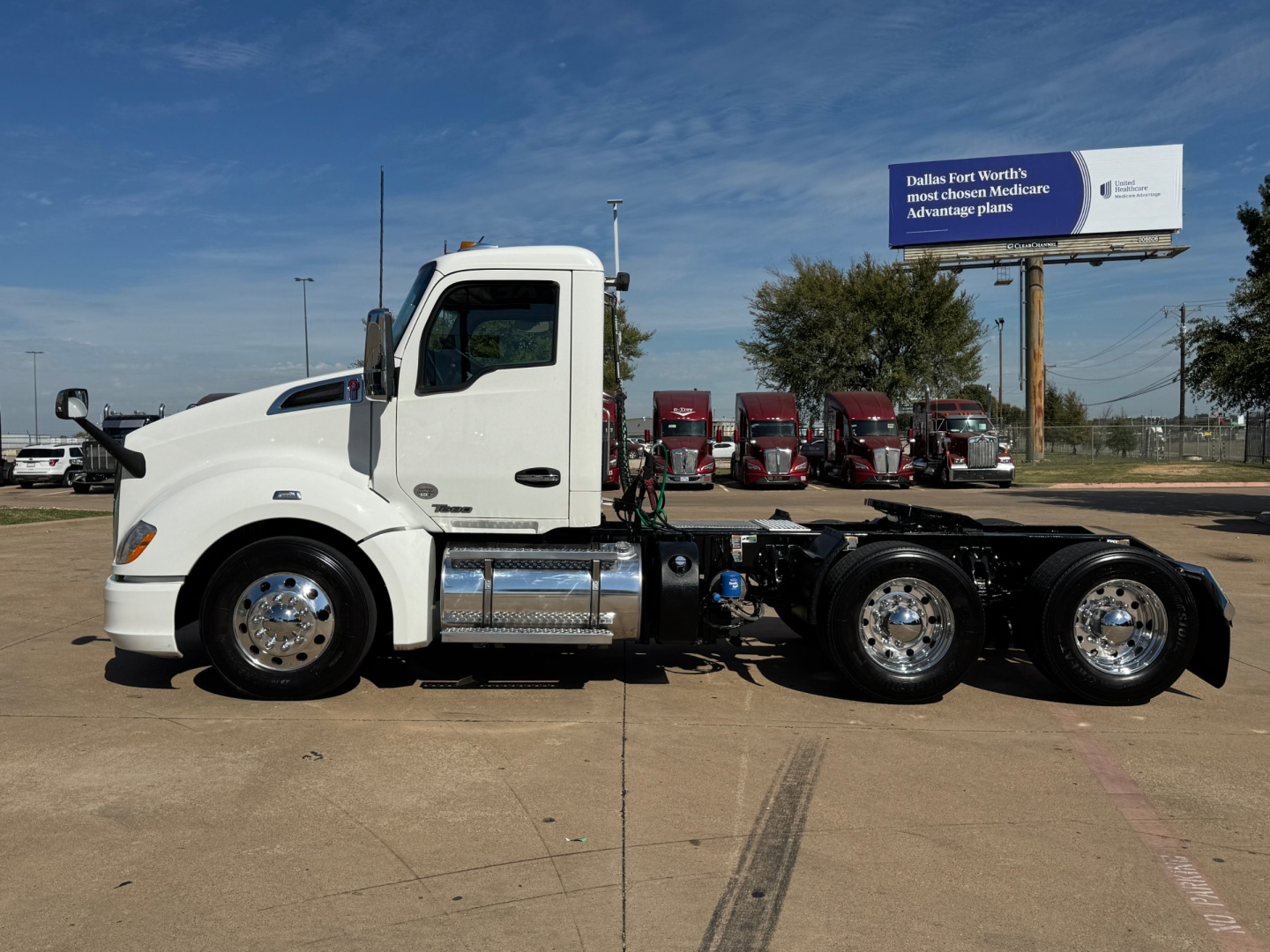 Kenworth T680 conventional daycab tractor, side view showing chrome fuel tanks and dual rear axles, white cab, billboards in background.