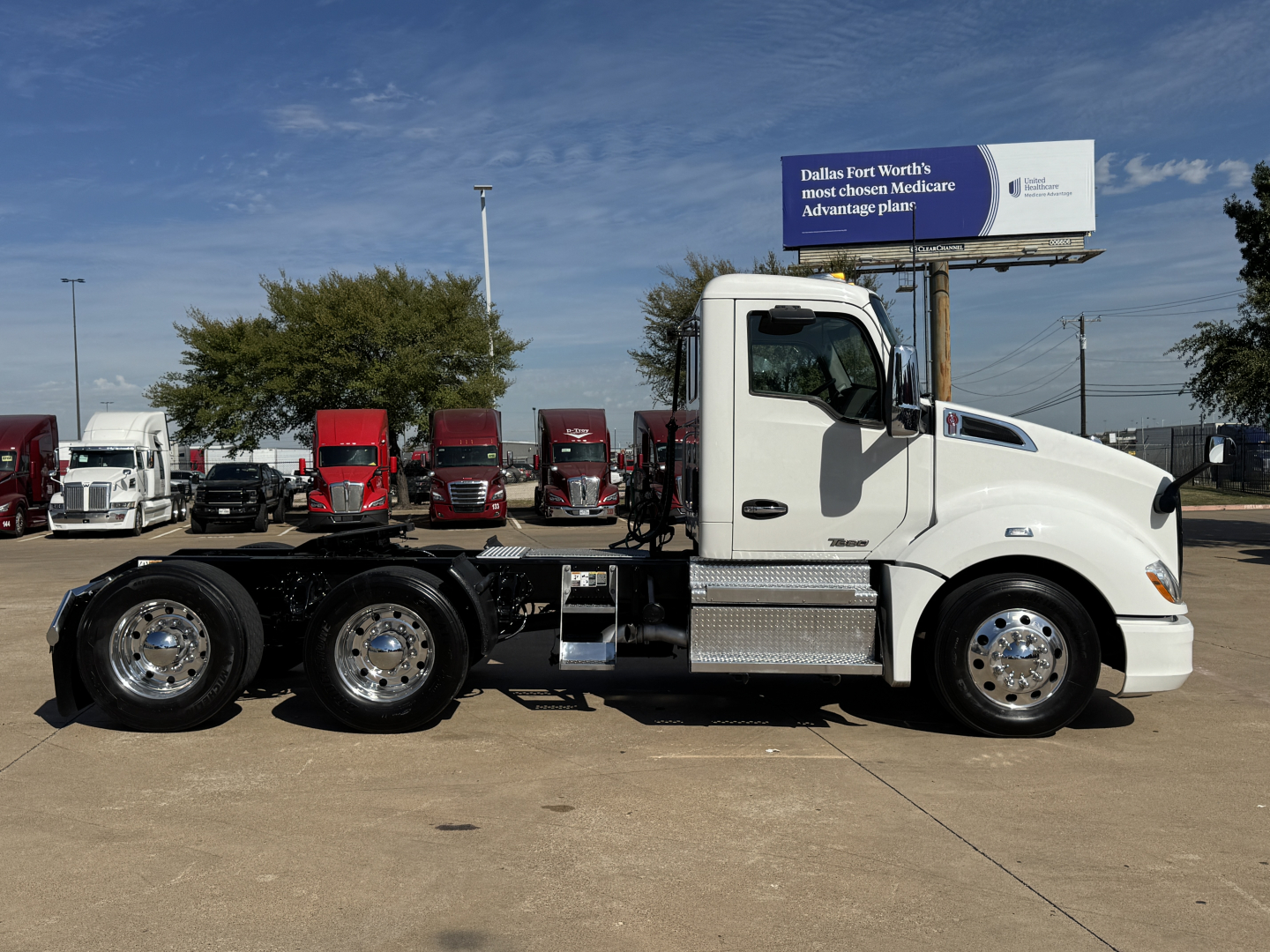 Kenworth T680 conventional daycab tractor, white with chrome wheels, side view showing fuel tanks and dual rear axles; background features signboard and other trucks.