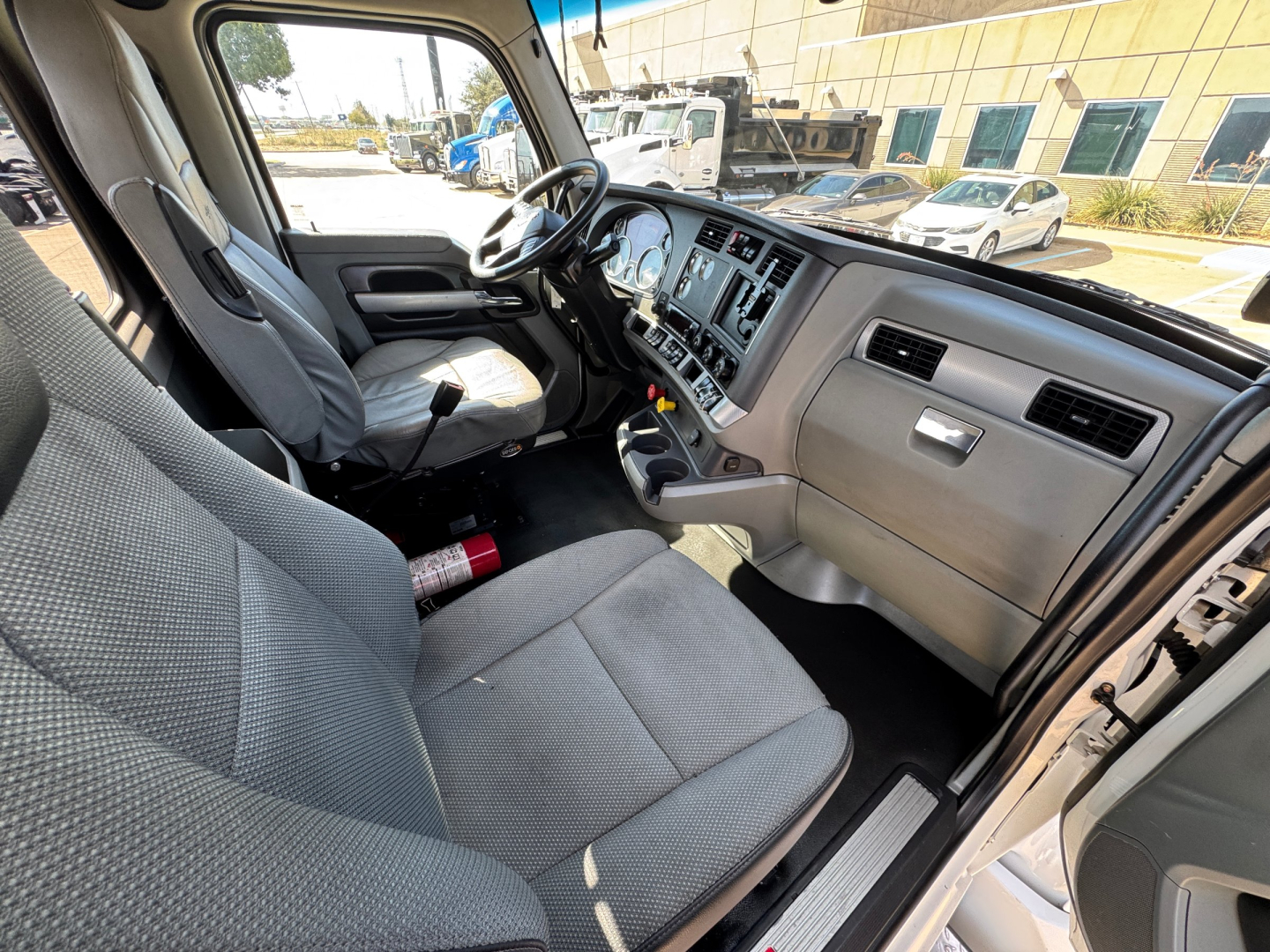 Kenworth T680 interior cab showing dash with multiple gauges and center console; gray fabric seats; steering wheel; open door; steering wheel and driver seat view.