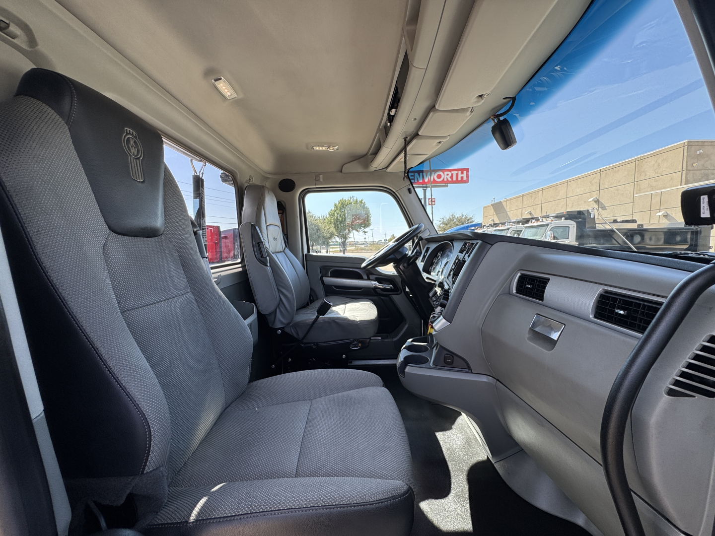 Cab interior of 2022 Kenworth T680 with front seats, dashboard, and steering wheel visible; driver and passenger seats cloth upholstery; overhead lighting.