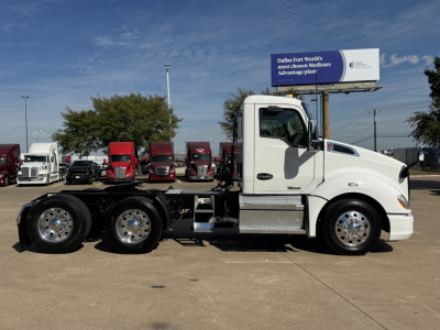 Kenworth T680 conventional daycab tractor, white with chrome wheels, side view showing fuel tanks and dual rear axles; background features signboard and other trucks.