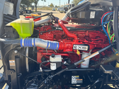 Kenworth T680 diesel engine bay with red engine, blue intake hose, yellow coolant reservoir, and various hoses and labels visible.