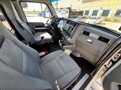 Kenworth T680 interior cab showing dash with multiple gauges and center console; gray fabric seats; steering wheel; open door; steering wheel and driver seat view.