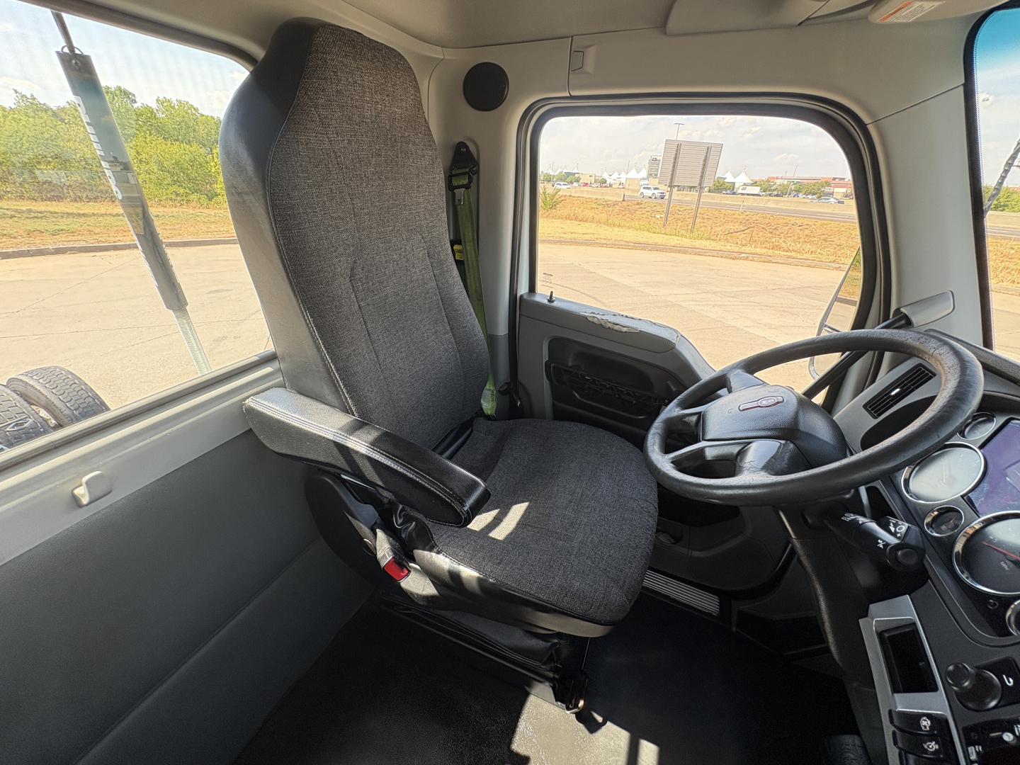 Kenworth T880 cab interior showing steering wheel and driver seat from a front-right perspective; dashboard gauges visible; gray fabric seat; window view of an outdoor yard