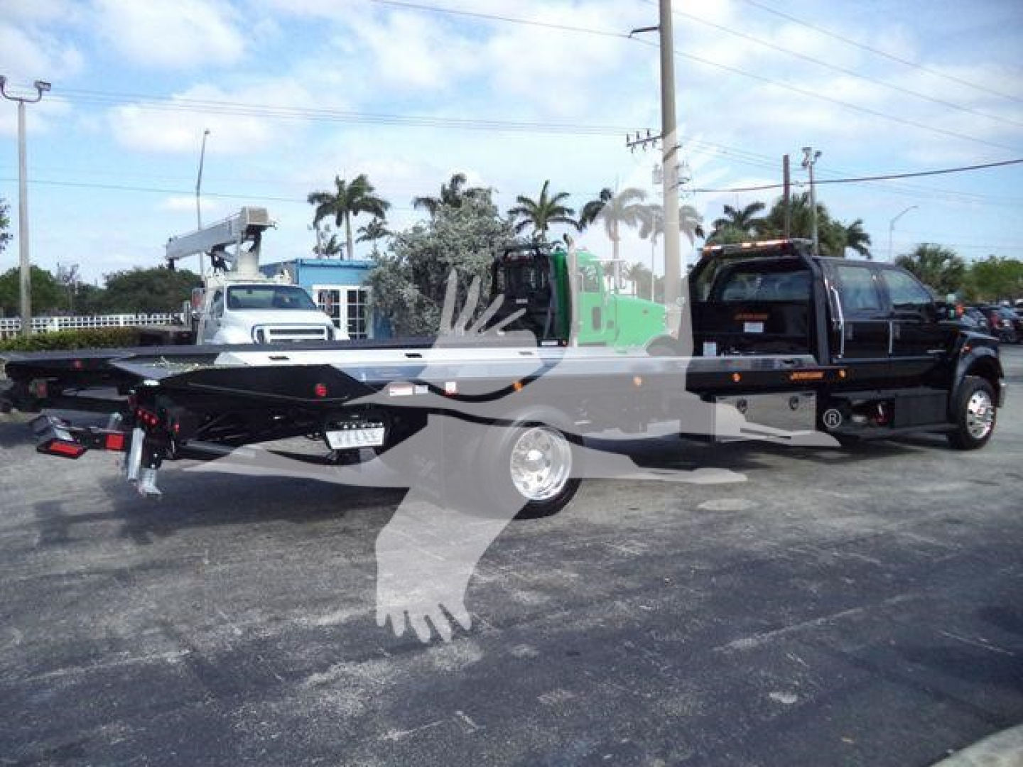 Ford F750 rollback tow truck side view showing flatbed and cab, black truck with green equipment in background, dealership lot with palm trees