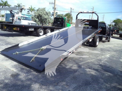 Flatbed tow truck with a long steel ramp extending from the chassis. Dual rear axles, black cab, and yellow safety cones. Visible on the truck are metal hooks and tie-down chains on the ramp.