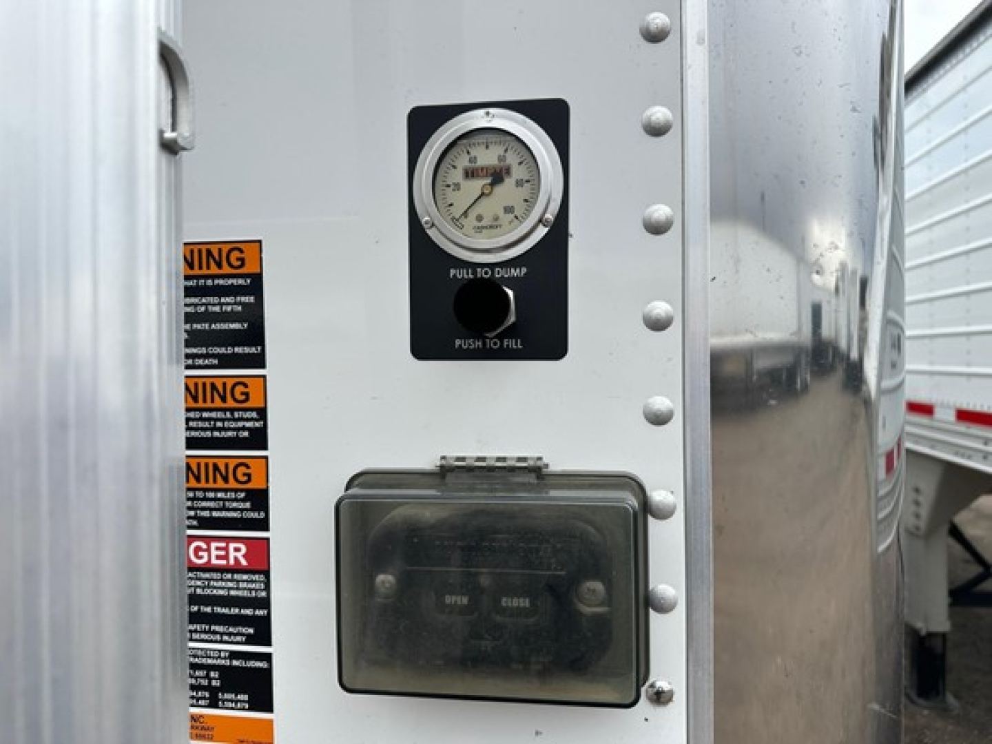 Trailer rear panel with pressure gauge labeled 'PULL TO DUMP PUSH TO FILL' and a rectangular black control box; warning stickers on left edge of aluminum surface.