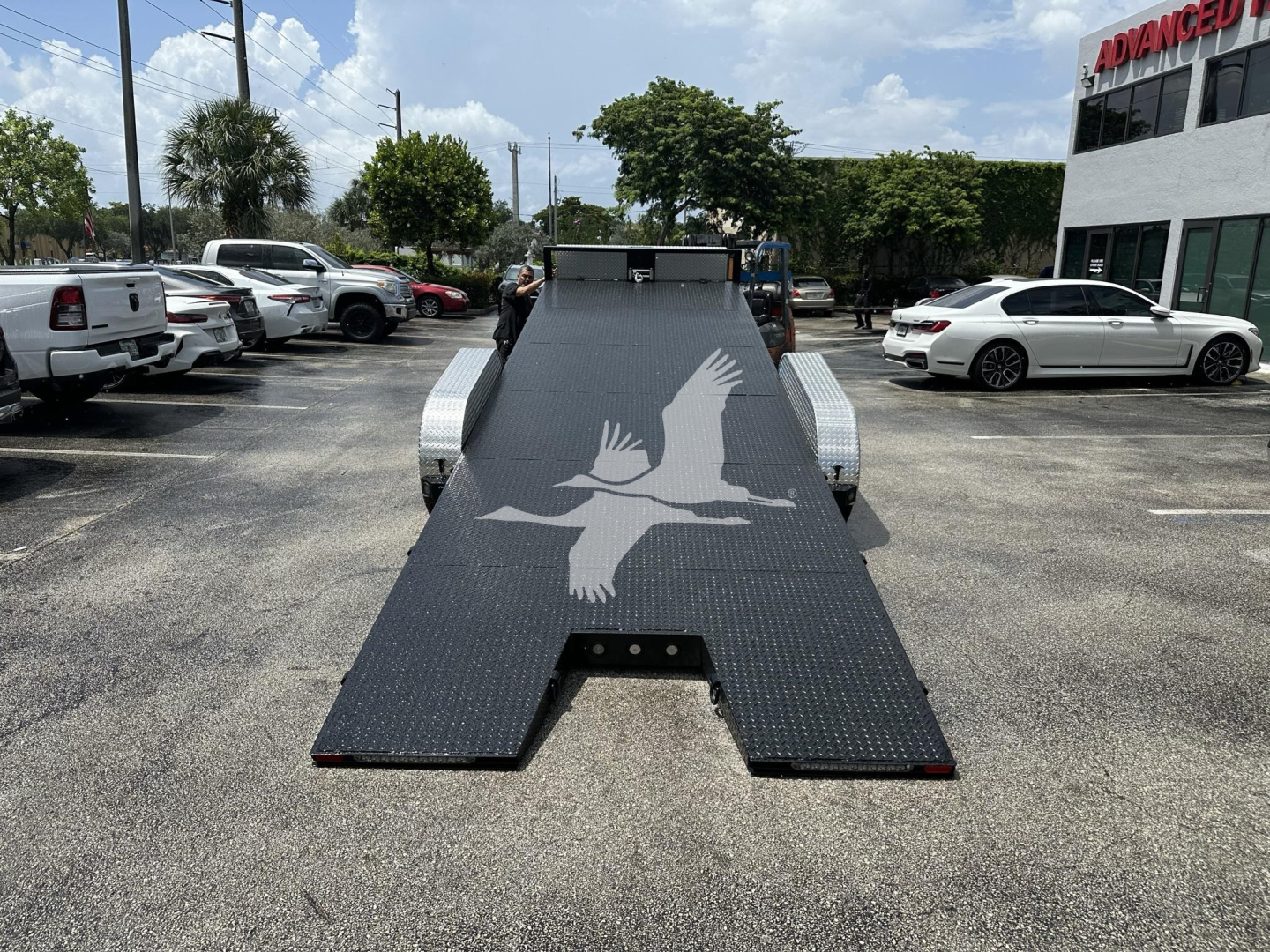 Flatbed tow trailer ramp with a diamond plate surface and a large white bird silhouette logo, parked in a dealership lot. Side fenders, wheel wells, and surrounding vehicles visible.