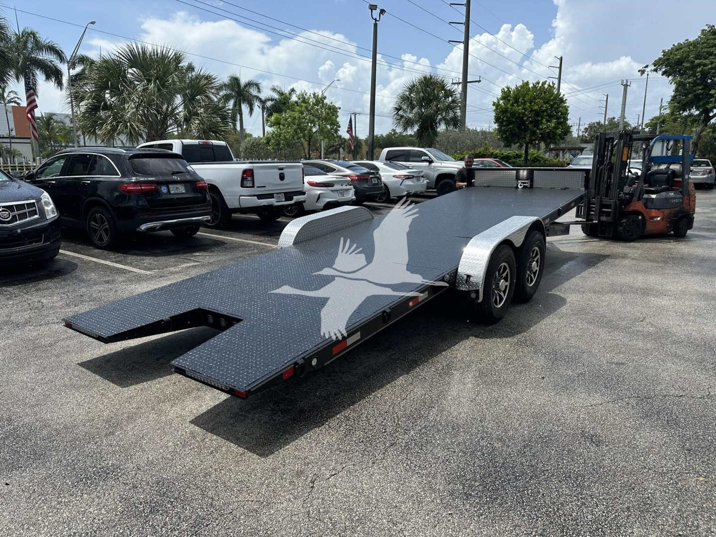 Flatbed trailer with dual axles and extended loading ramps in a parking lot, forklift visible on the right, palm trees and vehicles in background.