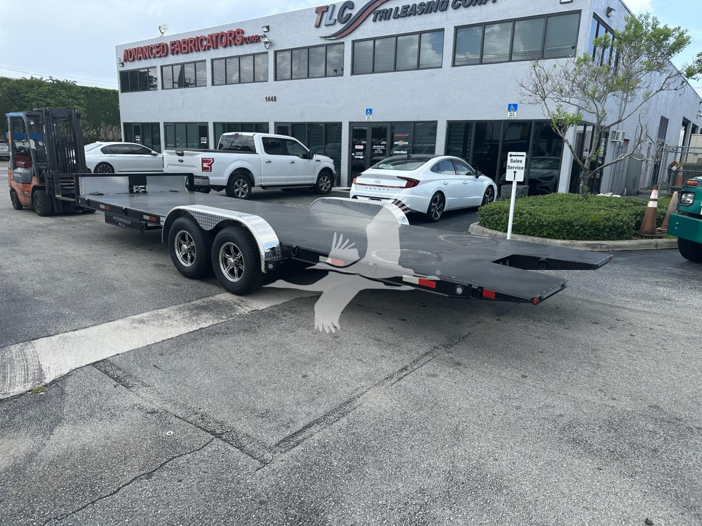 Trailer flatbed with dual axles and ramp extending, parked in lot outside TLC Leasing & General at a two-story showroom building with several cars around.