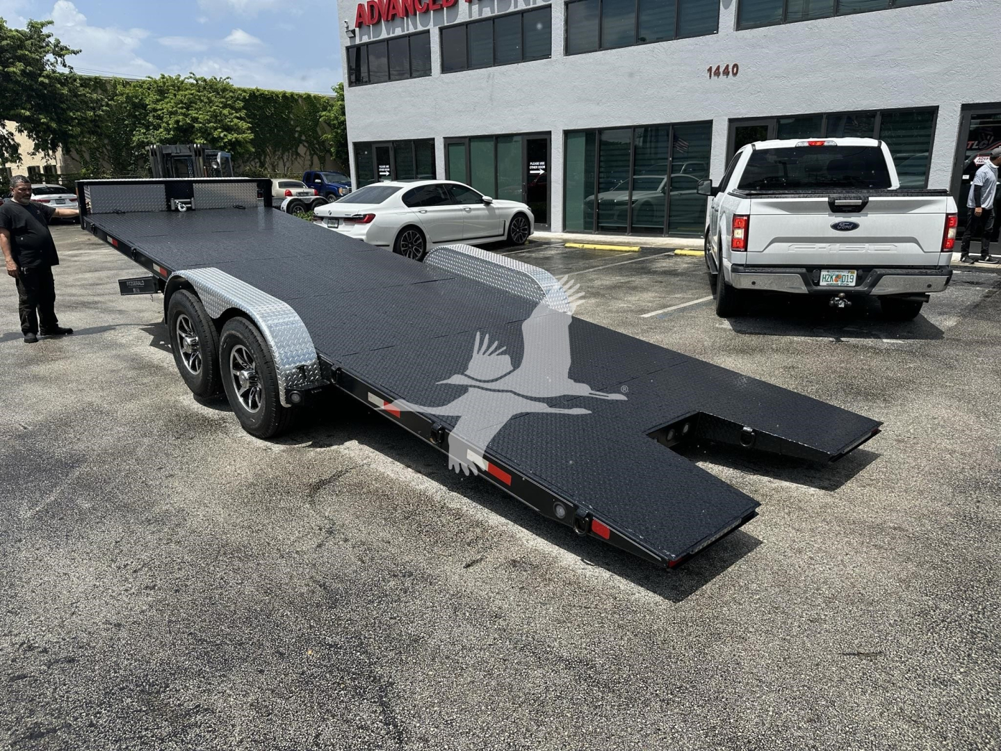 Flatbed trailer with dual axles and ramp, black steel deck, parked in front of a building with a white SUV nearby.