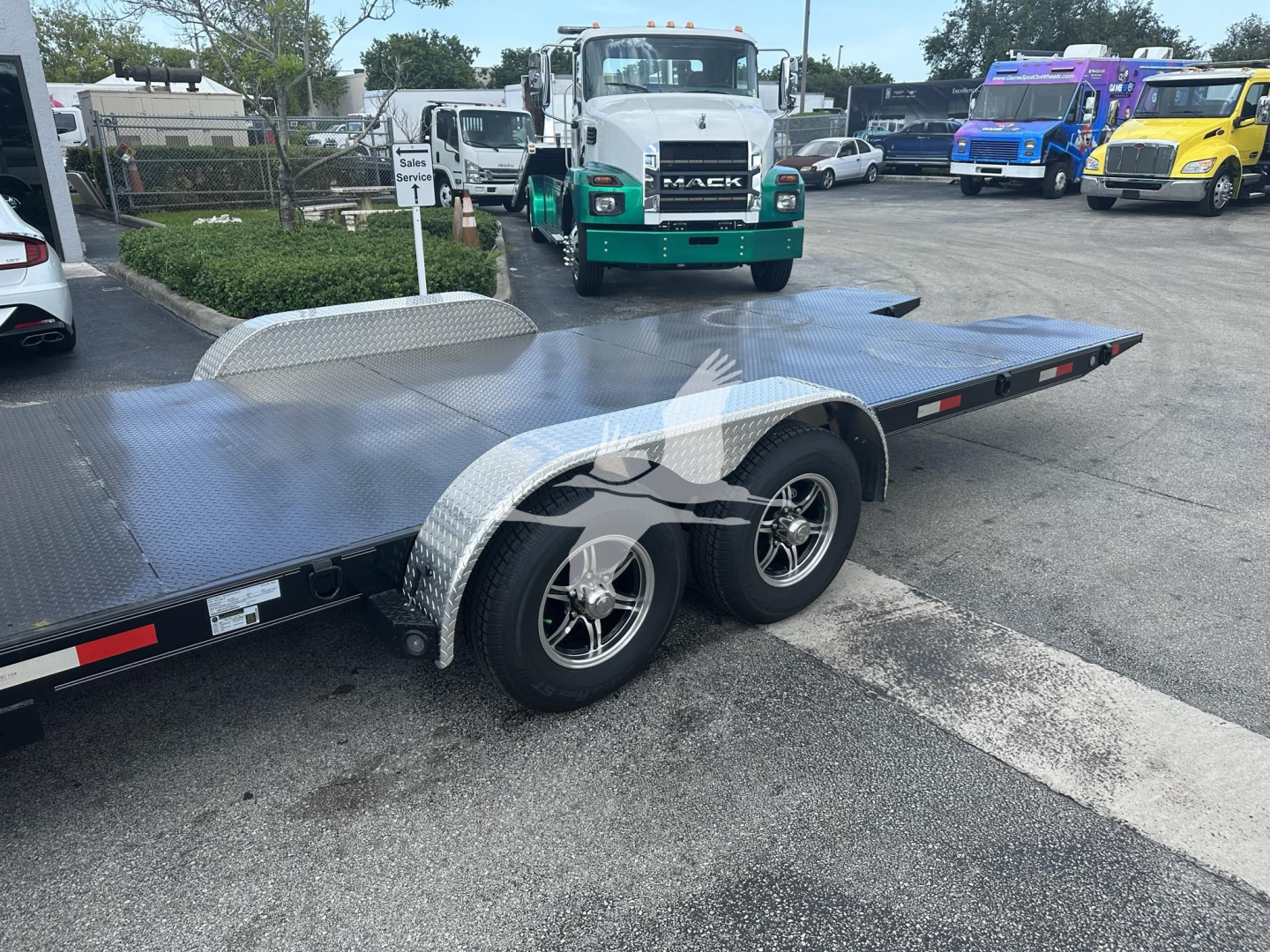 Flatbed trailer with dual wheels and aluminum diamond plate deck, fender, and side rails; adjacent green Mack truck in lot.