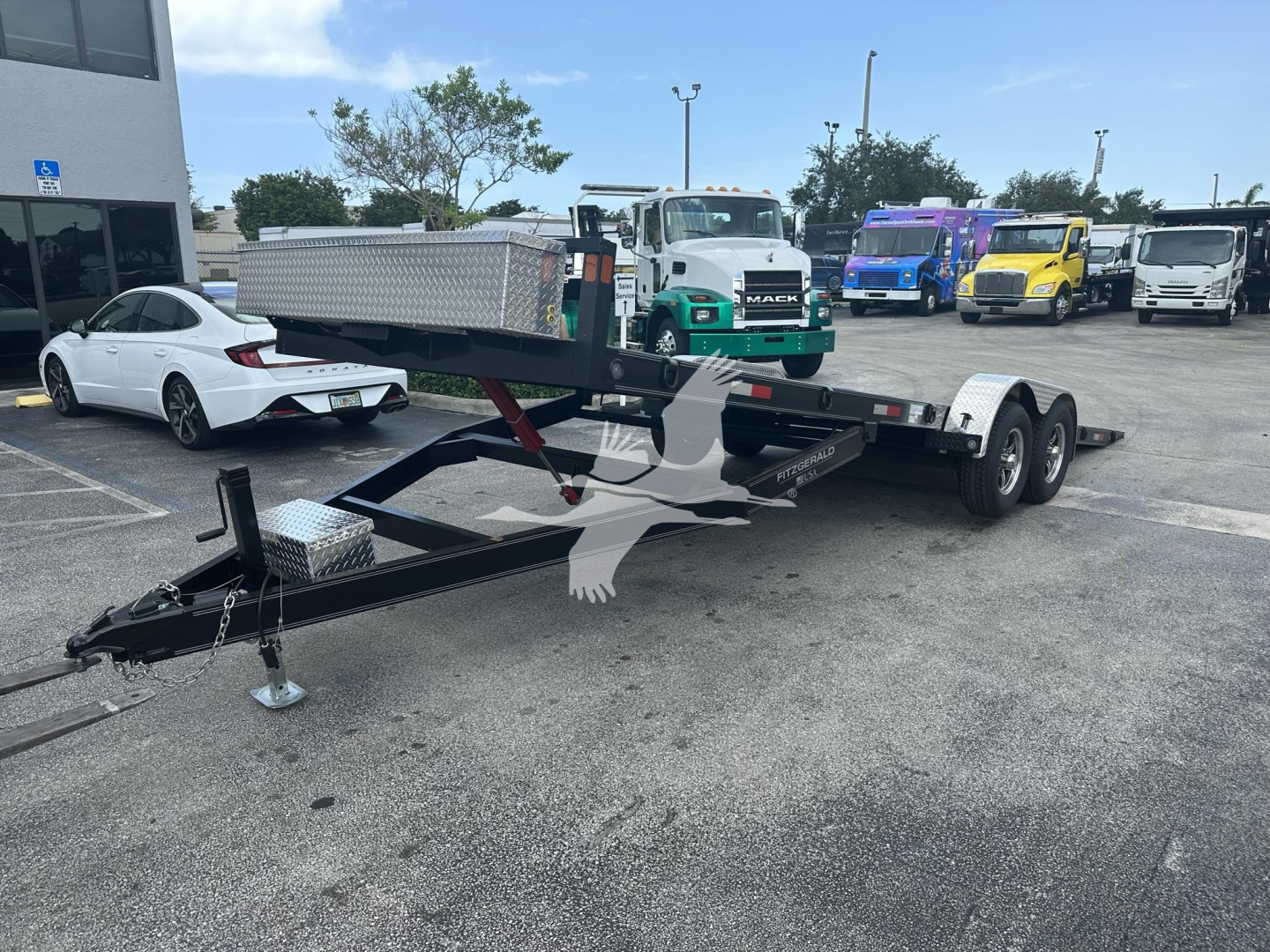 Trailer showing dual-axle equipment trailer with black frame and gooseneck tongue, safety chains, and diamond-plate toolbox. Overhead: other trucks and a white car in a lot.