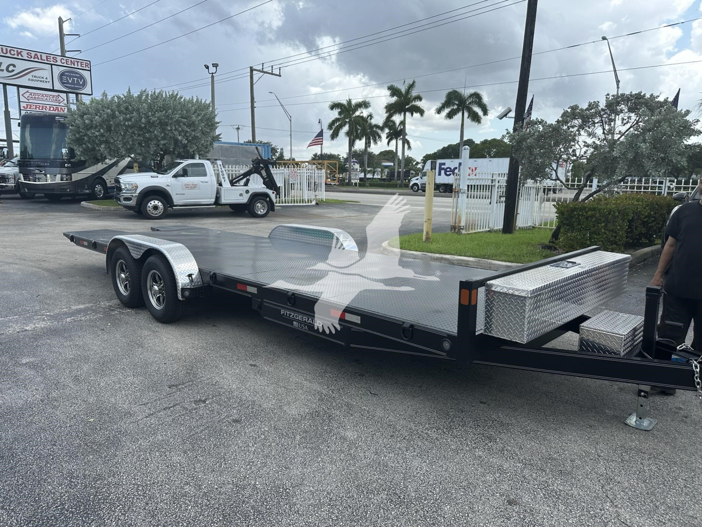 Equipment trailer with dual axles and a long flatbed, aluminum tool boxes on the sides, diamond plate storage compartments, and a gooseneck hitch area visible.