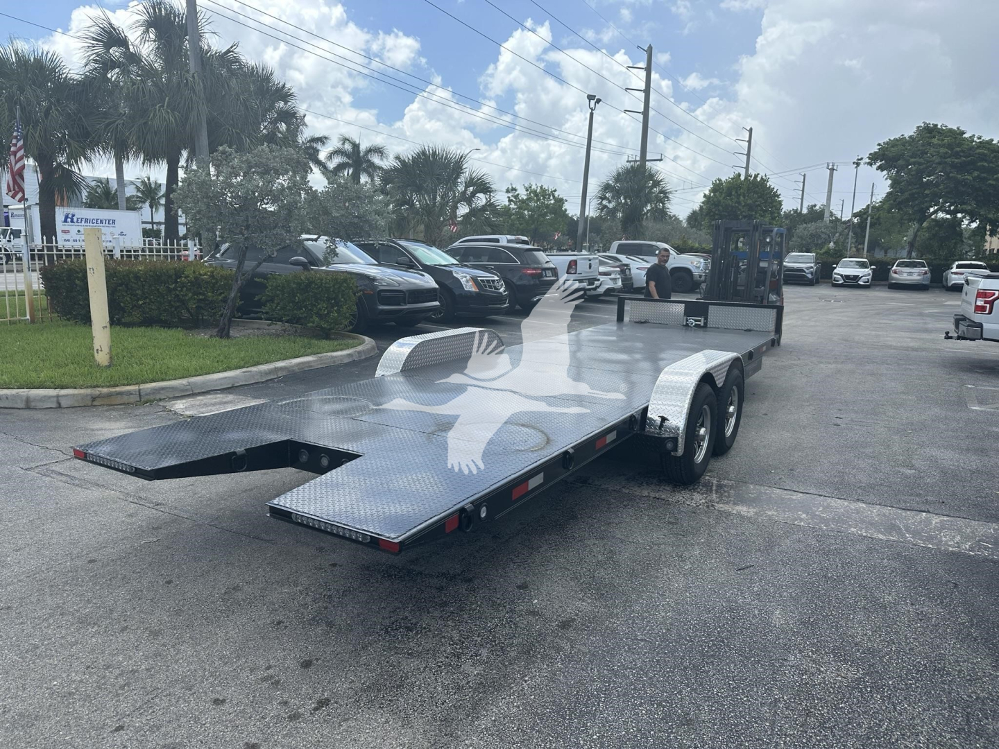 Trailer in a dealership lot with a flatbed car hauler and a ramp lowered, dual-axle trailer, metallic diamond-plate surface, storage area at the front, several vehicles in background.