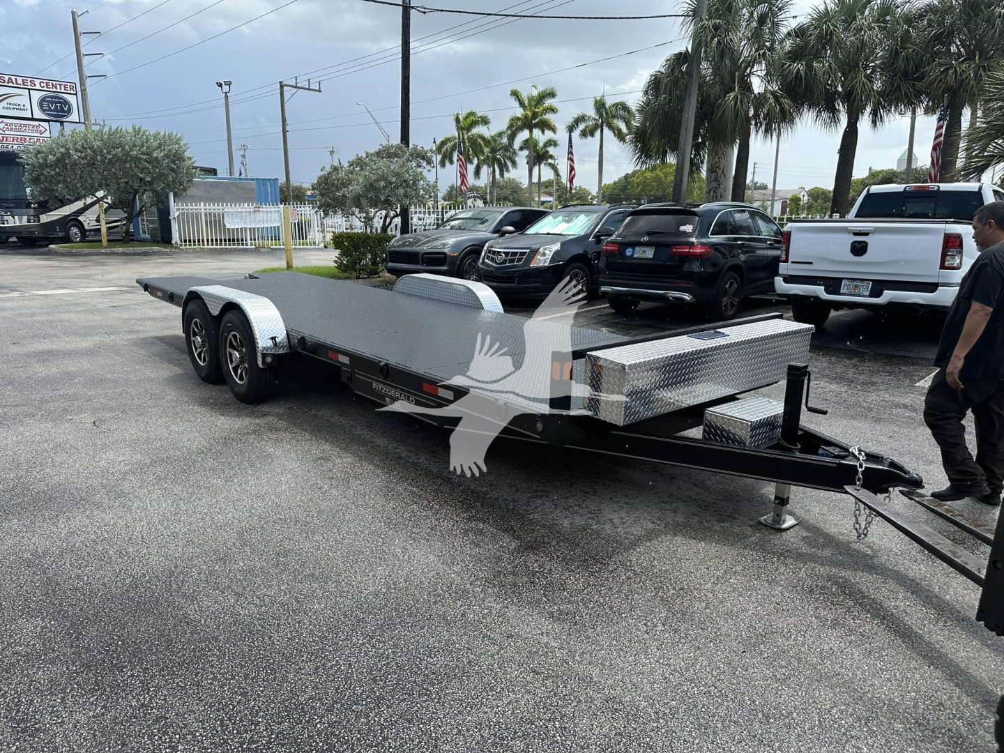 Flatbed trailer with dual axles and a diamond plate front, hitch at the front, showing a metal toolbox and support stands; some vehicles and palm trees in the background.