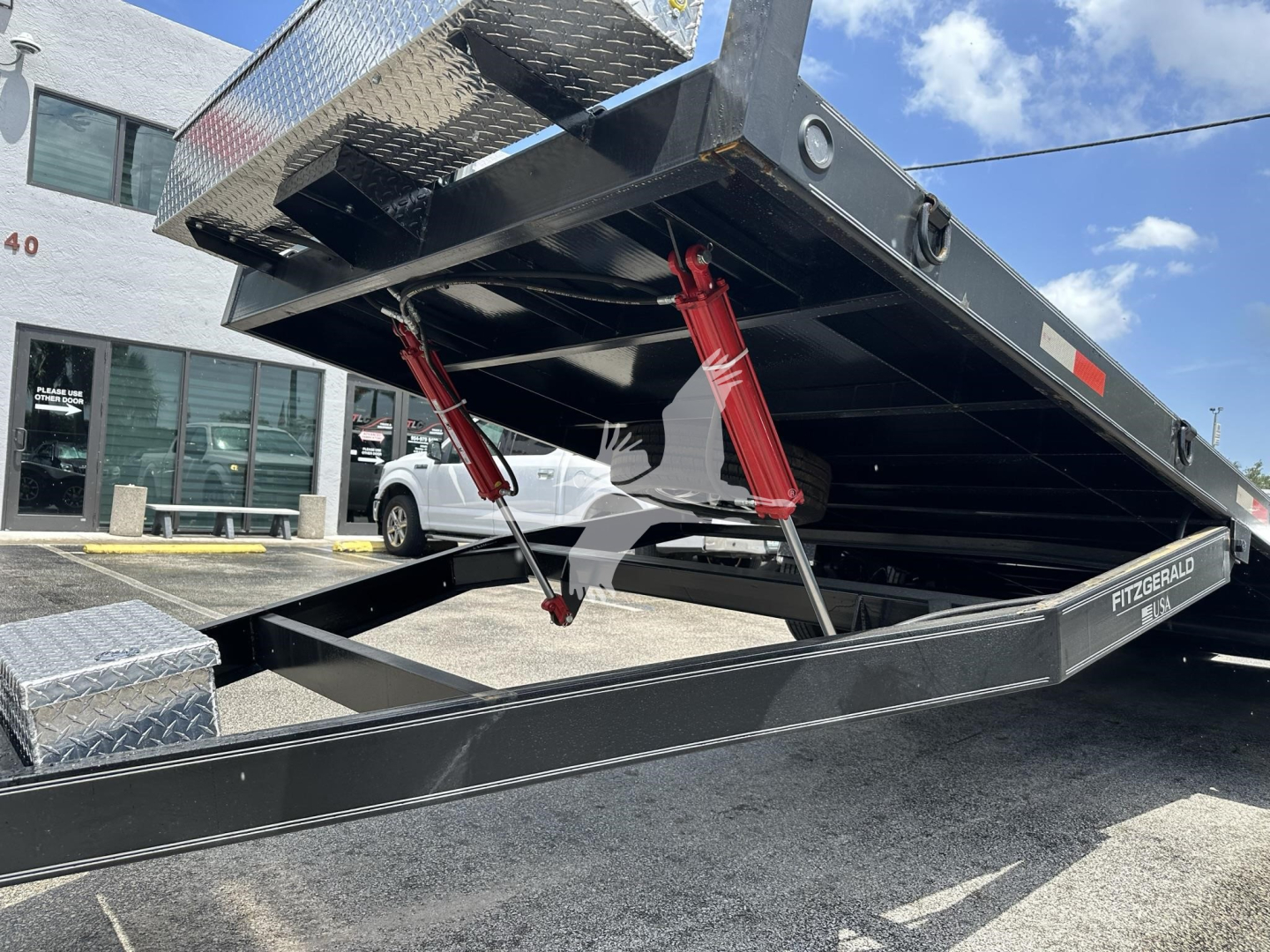 Trailer bed with hydraulic scissor jacks and tilt mechanism in a lowered position, black metal frame, diamond plate toolbox visible, outdoor lot.