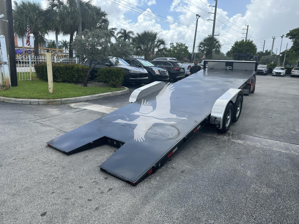 Trailer with a lowered ramp on a flatbed, dual-axle with aluminum side rails, gray surface with graphic; parked lot with other vehicles in background.