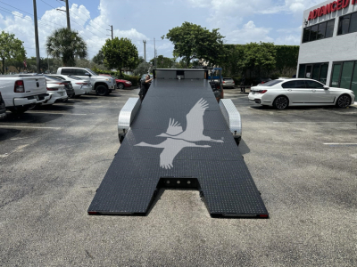 Flatbed tow trailer ramp with a diamond plate surface and a large white bird silhouette logo, parked in a dealership lot. Side fenders, wheel wells, and surrounding vehicles visible.