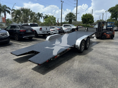 Flatbed trailer with dual axles and extended loading ramps in a parking lot, forklift visible on the right, palm trees and vehicles in background.