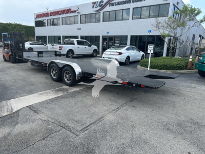 Trailer flatbed with dual axles and ramp extending, parked in lot outside TLC Leasing & General at a two-story showroom building with several cars around.