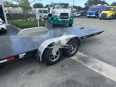 Flatbed trailer with dual wheels and aluminum diamond plate deck, fender, and side rails; adjacent green Mack truck in lot.