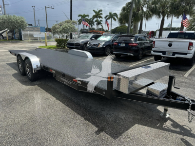 Trailer-style equipment trailer with dual axles and diamond plate ramp in a parking lot.