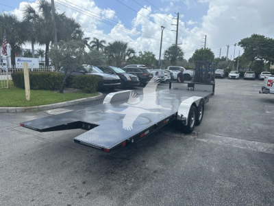 Trailer in a dealership lot with a flatbed car hauler and a ramp lowered, dual-axle trailer, metallic diamond-plate surface, storage area at the front, several vehicles in background.